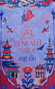 Das Cover des Romans "The Girl Who Fell Beneath The Sea" von Axie Oh zeigt die stilisierte Darstellung einer asiatischen Stadt, die sich scheinbar unterhalb des Meeres befindet, sowie eines herabsinkenden Mädchens unterhalb der Meeresoberfläche.